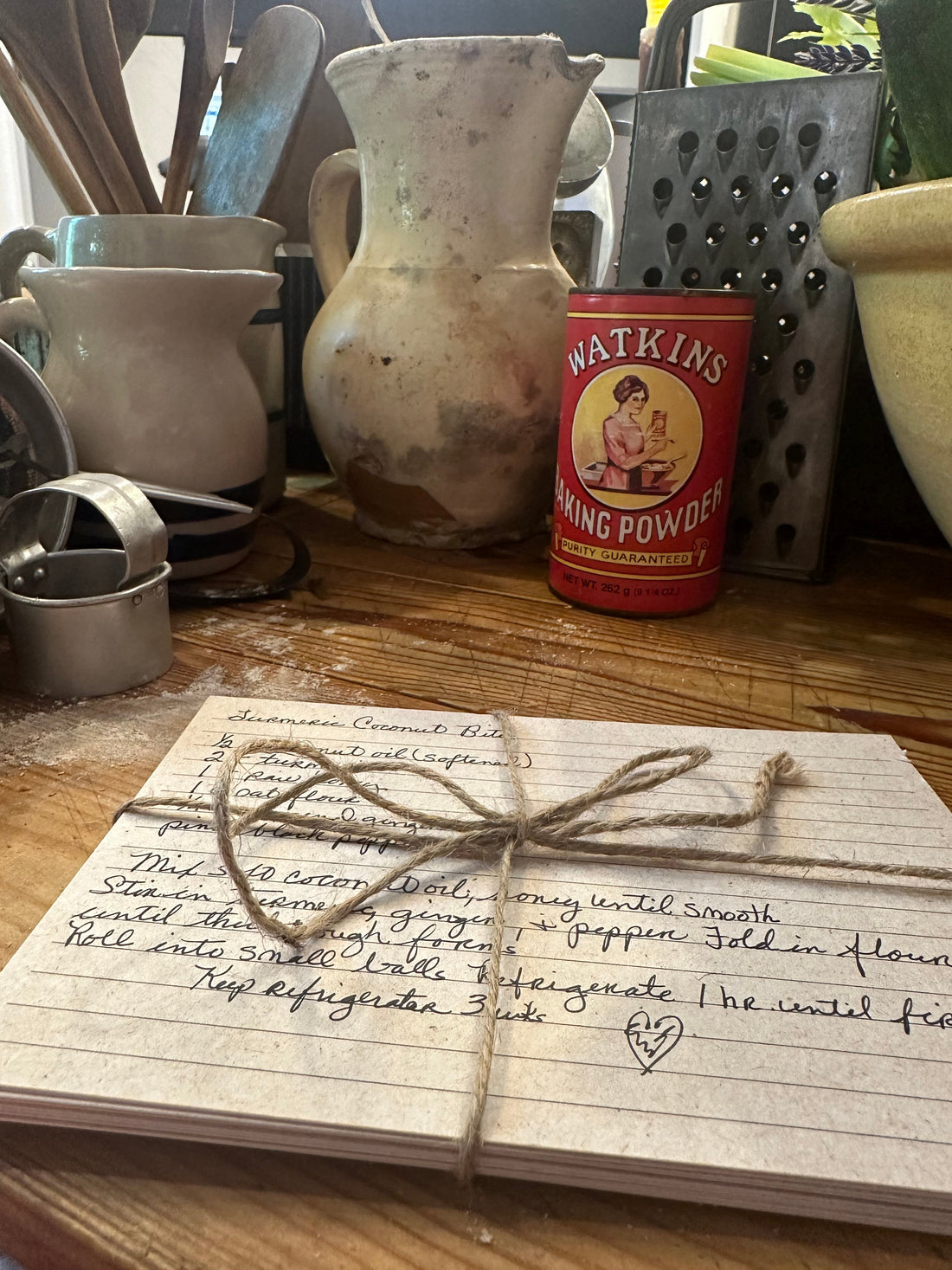 Handwritten recipe cards tied with twine next to a vintage Watkins baking powder tin and well-loved kitchen tools" 