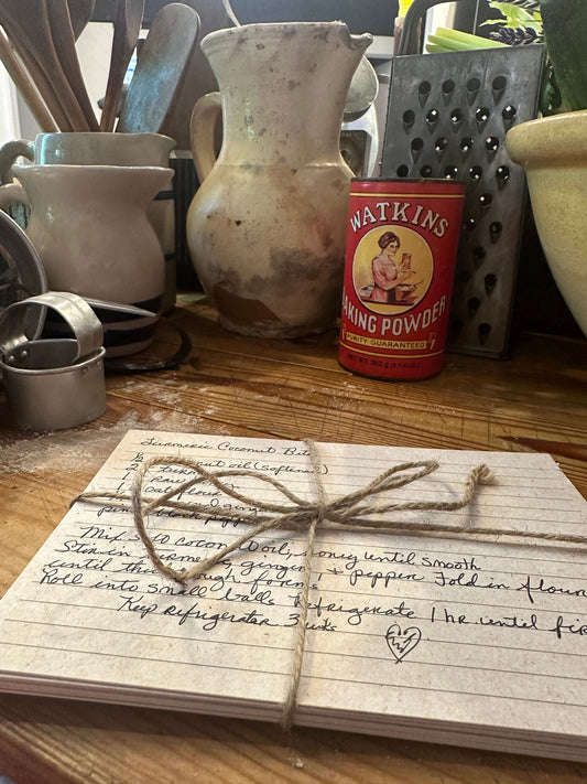 Handwritten recipe cards tied with twine next to a vintage Watkins baking powder tin and well-loved kitchen tools" 
