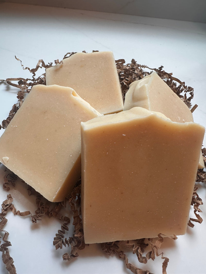 Four rectangular bars of pale beige soap arranged on brown shredded paper against a white background.