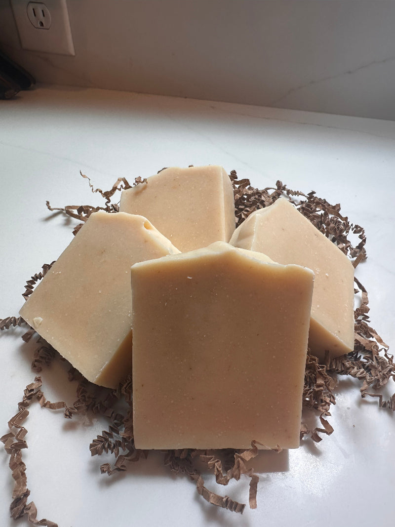 Four rectangular bars of handmade soap in a warm light tan color, arranged on a bed of brown crinkle paper shred on a white surface.