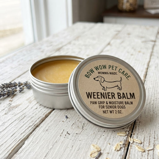 An open round metal tin of Weenier Balm pet balm sits on a rustic white wooden table, with the lid leaning in front. The lid label reads 'BOW WOW PET CARE' and shows a dachshund illustration.